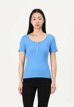Modrý žebrovaný top s krátkými rukávy a kulatým výstřihem, se zapínáním na knoflíky na hrudi. Nosené s černými skinny kalhotami.