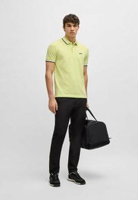 Ljusgul polo med svart och vit krage, i kombination med svarta byxor och en svart väska. Outfitten inkluderar svarta sneakers med vita detaljer.