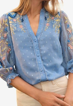 Frau trägt eine blaue transparente Bluse mit floraler Stickerei an Ärmeln und Schultern, vorne mit Knöpfen, kombiniert mit beigen Hosen.