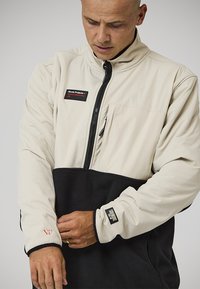 Giacca a pullover con parte superiore color crema e fondo in pile nero, dotata di mezza zip, tasca sul petto e dettagli con logo in rosso e nero.