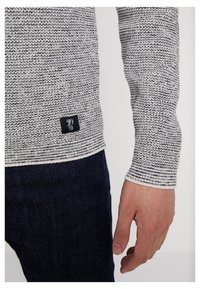 Gestrickter Pullover in Grau und Weiß mit einem strukturierten Muster, versehen mit einem schwarzen Etikett am Saum. Kombiniert mit dunkelblauen Jeans.