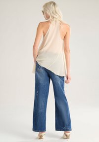 Top beige senza maniche con orlo asimmetrico, abbinato a jeans blu a gamba larga con texture strappata e lavaggio sbiadito.