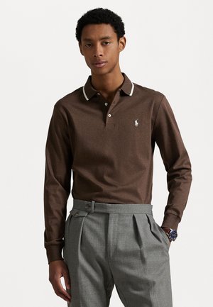Bruine langemouwen polo met witte kraagaccenten en een klein logo op de borst. Gecombineerd met lichtgrijze high-waisted broek.
