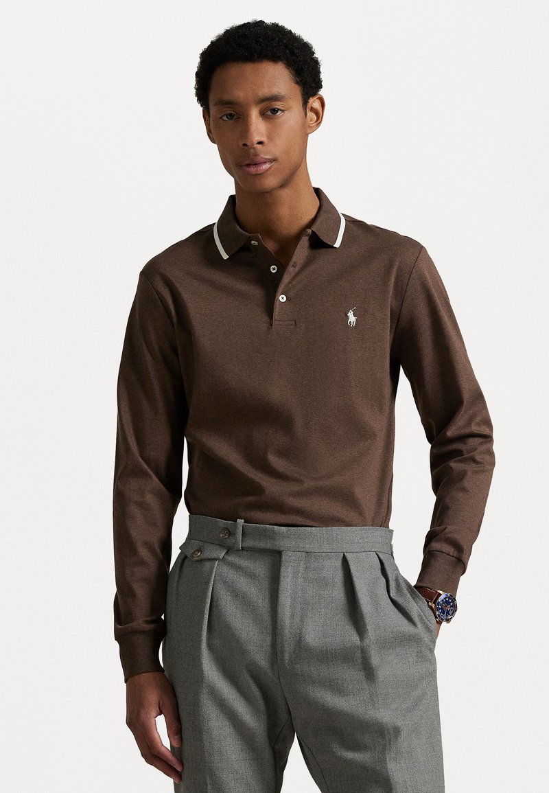 Bruine langemouwen polo met witte kraagaccenten en een klein logo op de borst. Gecombineerd met lichtgrijze high-waisted broek.