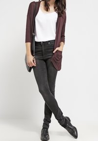 Žena stojící s překříženýma nohama, oblečená v černých džínách, bílém tričku, tmavě fialovém kardiganu, černých botách a s černou kabelkou na rameni.