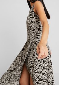 Kevyt mekko ruskeassa kangassa, jossa on mustia pilkkuja. Omistaa hihattoman muotoilun, tarjoilee virtaavaa helmaa ja sivureiän lisäämään liikettä.