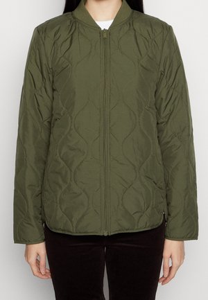 Frau trägt eine olivgrüne gesteppte Jacke mit Frontreißverschluss, Rippkragen und langen Ärmeln, kombiniert mit dunkler Hose.