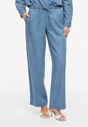 Nő laza, világoskék farmernadrágban, gumis, zsinóros derékrésszel és fehér, nyitott orrú magassarkú szandálban, fehér háttér előtt áll.