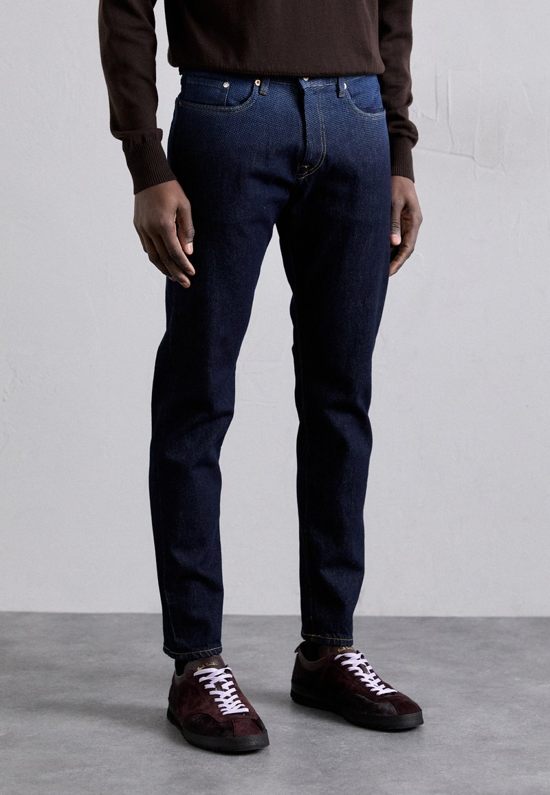 Jeans de denim azul oscuro con un corte ajustado, que presentan un diseño clásico de cinco bolsillos y remaches de cobre. Combinados con zapatos burdeos y un suéter marrón.