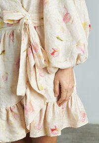 Blumenkleid aus leicht beigem Stoff, mit rosa und gelben Blumenmustern, voluminösen Ärmeln, einem Bindegürtel und Rüschensaum.
