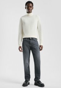 Młody mężczyzna stoi, ubrany w biały sweter z golfem, ciemnoszare dżinsy i czarne, masywne buty na tle jednolitego jasnego tła.