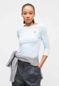 Γυναίκα με πλεξούδες, φοράει πουκάμισο με ανοιχτό μπλε και λευκές ρίγες και μικρό έμβλημα φράουλας, σκούρο παντελόνι και γκρι πουλόβερ δεμένο γύρω από τη μέση.