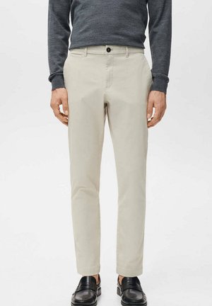 Uomo che indossa pantaloni beige chiaro, maglione scuro e scarpe eleganti nere in piedi contro uno sfondo bianco semplice, mani lungo i fianchi.