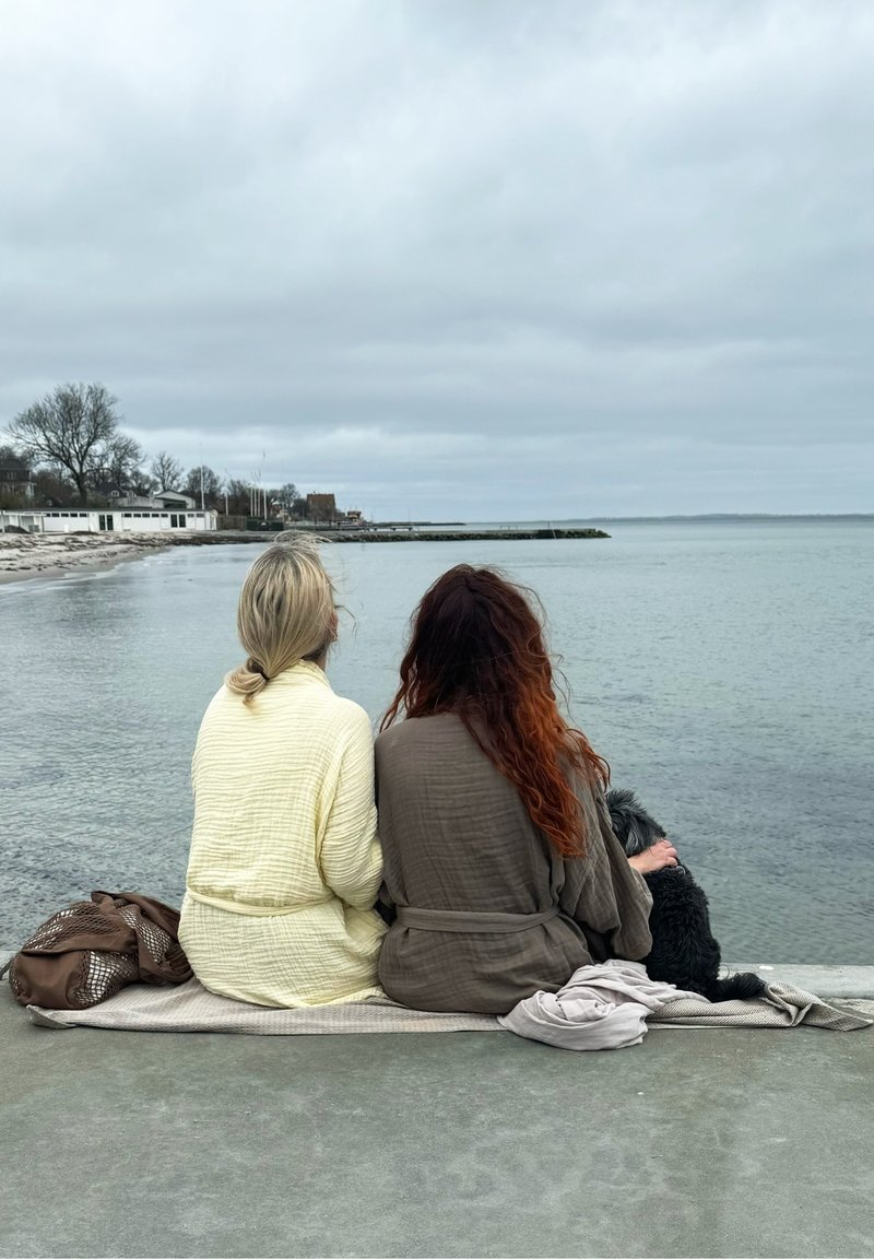 Divi cilvēki sēž uz segas pie ūdens, ģērbušies teksturētās, gaiši dzeltenās un pelēkās drēbēs. Tumšs suns guļ blakus viņiem.