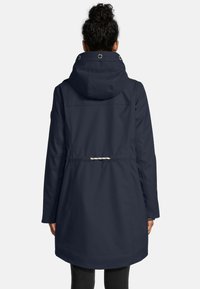 Vrouw in een marineblauwe mid-length regenjas met capuchon en een getailleerde taille, gezien van achteren tegen een effen achtergrond.