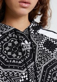 Černá mikina s kapucí s bílými geometrickými a paisley vzory. Nastavitelné šňůrky na kapuci a vyšívané logo na hrudi.