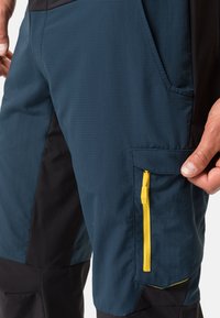 Shorts multi-panneaux bleu et noir avec un tissu texturé, poche latérale avec fermeture éclair jaune et un design ajusté pour un maintien sécurisé.