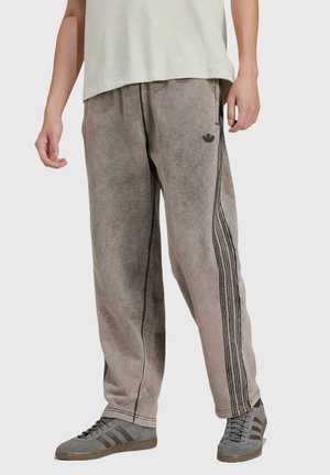 Pantaloni in morbido tessuto marrone con vestibilità ampia, caratterizzati da strisce laterali nere e un logo ricamato. Indossati con scarpe grigie.