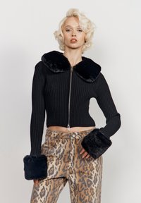 Juodas gumbuotis trumpas švarkas su pūkuota apykakle ir rankovėmis, derinamas su leopardų rašto plačiomis kelnėmis su kišenėmis ir siuvinėjimo detalėmis.