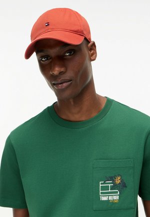 Hombre que lleva una gorra de béisbol coral y una camiseta verde con un bolsillo delantero que presenta un logo floral de Tommy Hilfiger.