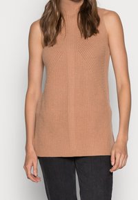 Nainen pukeutuneena hihattomaan ruskeaan neulottuun pooloneuleeseen ja mustiin housuihin seisoo yksinkertaisen vaalean taustan edessä.