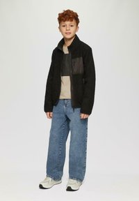 Zwarte fleecejack met textuurstof en een zwart ritssluiting borstzak. Gecombineerd met lichtblauwe jeans en witte sneakers.