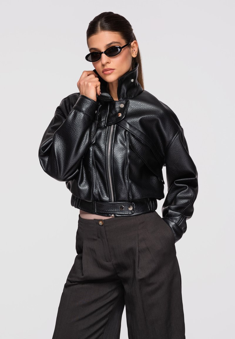 Femme portant une veste courte en cuir noir, un pantalon large foncé, et de petites lunettes de soleil noires, posant avec une main près du visage.