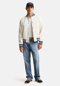 Young man wearing a white varsity jacket with navy stripes, blue jeans, white polo shirt, black sneakers, and a navy cap with US flag embroidery.