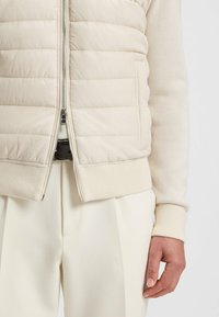 Hellbeige Steppweste mit horizontaler Quiltoptik, Reißverschluss und gerippten Strickbündchen. Kombiniert mit passenden hellbeigen plissierten Hosen.