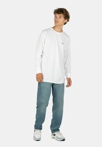 Camisa de algodón blanca de manga larga con cuello redondo, con un sutil logotipo en el pecho, combinada con jeans azul claro y zapatillas blancas.