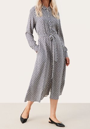 Robe à manches longues en tissu léger avec un motif de feuilles bleu et blanc, taille cintrée avec un lien, et jupe longueur cheville. Chaussures plates noires.