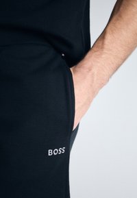 Îmbrăcăminte sport neagră, realizată dintr-un material texturat, cu un buzunar lateral și un logo "BOSS" brodat discret în alb.