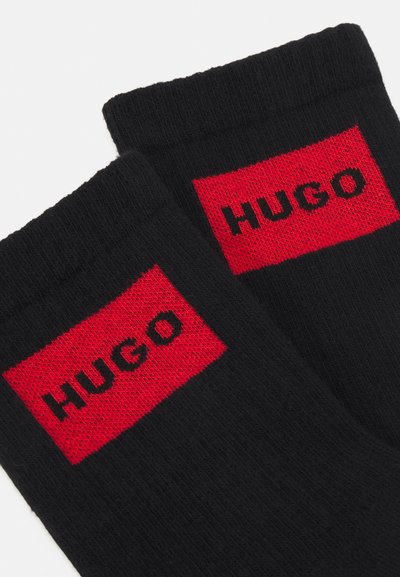 Chaussettes noires avec une texture ribbed, ornées d'un patch rectangulaire rouge brodé avec "HUGO" en noir.