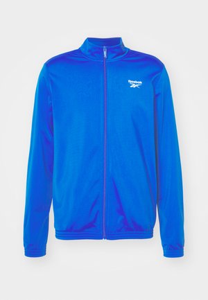 Blå zip-up jacka i lättviktsmaterial. Har en hög krage och elastiska ärmslut. Logotyp på vänster bröst. Slät textur.