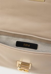 Bē�šas ādas rokassomas iekšpuse ar zelta krāsas aizdari, melnu logotipa birku un gaišu auduma oderi ar smalku rakstu.