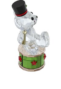 Kryštáľová figúrka medveďa s čiernym cylindrom, sediaca na zelenej bubne zdobenej červenými hviezdami a zlatou paličkou na bubne. Priesvitná textúra.