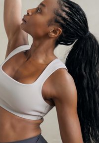 Top corto deportivo gris claro con tirantes anchos, diseño ajustado y textura suave; cabello negro largo trenzado estilizado en una coleta.
