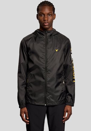 Jonge man met een zwart jack met rits en capuchon, met een geel logo op de borst en gele tekst op de rechtermouw tegen een grijze achtergrond.