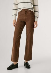 Bruine corduroybroek met een uitlopende ontwerp. De stof heeft verticale ribbels en is gecombineerd met een crèmekleurige gestreepte sweater en zwarte schoenen.