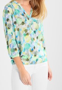 Lekka, przejrzysta bluzka z mieszanką zielonych, niebieskich i żółtych okrągłych wzorów. Dekolt w serek z ozdobnymi guzikami oraz rękawami o długości do łokcia.