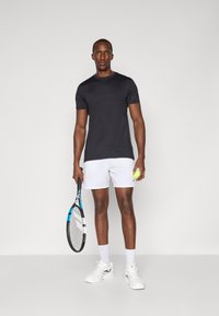 T-shirt de sport noir à manches courtes, shorts blancs, tenant une balle de tennis jaune et une raquette. Tissu texturé, design ajusté, chaussures de sport blanches.