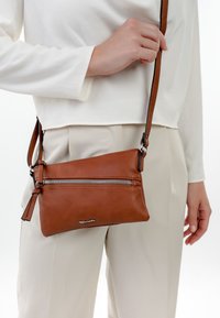 Sac bandoulière en cuir marron avec poche zippée à l'avant porté par une personne vêtue d'un haut à manches longues blanc et d'un pantalon beige clair sur un fond blanc.