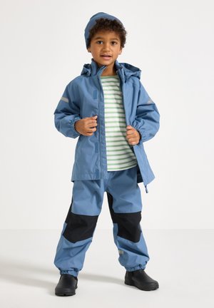 Barn iført en blå regnjakke og bukser med sorte knælapper, grøn- og hvidstribet skjorte, blå hue og sorte støvler, stående på hvid baggrund.