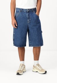 Shorts en denim, bleus, longueur mi-cuisse, coupe ample, design à cinq poches, ourlets cousus. Portés avec des chaussures de sport beiges et des chaussettes blanches.