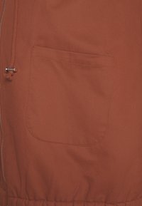 Tela de chaqueta color óxido con cremallera lateral y bolsillo parche expuesto. El material tiene una textura suave con detalle de cintura elástica.