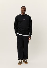 Άνδρας που στέκεται, φορώντας μαύρο φούτερ και αντίστοιχο μαύρο παντελόνι φόρμας με μπεζ αθλητικά παπούτσια, σε απλό φωτεινό φόντο.