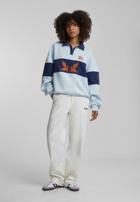 Lyseblå fleecegenser med marineblå striper, brodert oransje design og krage. Kombinert med hvite vidde bukser og joggesko.