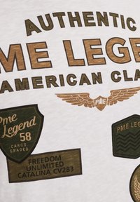 T-shirt blanc avec des patches imprimés marron et vert. Le texte inclut "One Legend 58," "Freedom Unlimited," et des détails de logo ailés.