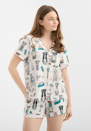 Vrouw draagt een korte mouw, knooppyjama met kleurrijke illustraties van honden en sportuitrusting op een lichte achtergrond.