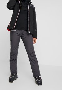 Schwarze Outdoor-Jacke mit einem gemusterten oberen Abschnitt, kombiniert mit grauen Hosen mit mehreren Taschen. Strukturiertes Gewebe, entworfen für kaltes Wetter.